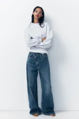Pantalón de jean celeste de tiro alto, corte wide leg y largo hasta el tobillo. Presenta un diseño asimétrico en la cintura con cierre frontal cruzado con cremallera y botones metálicos.