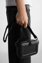 Bandolera rígida negra con formato de caja. Tiene un compartimento principal con cierre de cremallera, un bolsillo interior plano y asa de hombro ajustable y extraíble.