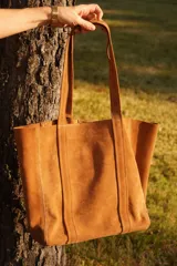 Cartera tote de gamuza color camel, con doble asa de hombro, bolsillo externo y detalle de borla colgante.
