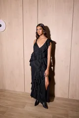 Vestido largo negro con escote en V y tirantes finos. Presenta volados verticales en cascada a lo largo del cuerpo y un tajo pronunciado en la parte delantera de la falda. La espalda es baja con tiras ajustables.