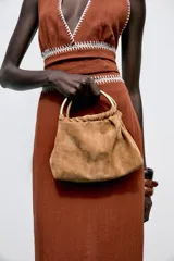 Bolso mini tipo saca de cuero serraje color marrón con efecto soft. Tiene doble asa de mano metálica dorada y asa bandolera extraíble. Cuenta con bolsillo interior con cremallera y cierre mediante fruncido y botón imantado.