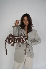 Bolso bandolera de cuero marrón con efecto desgastado y detalles pintados en blanco. Cuenta con dos bolsillos frontales con hebillas plateadas, correa de hombro ajustable y herrajes metálicos plateados.