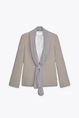 Blazer color beige con cuello solapa, manga larga con abertura y detalle de lazo gris en el frente.