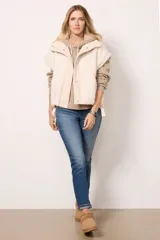 Chaleco de piel sintética tipo sherpa en color blanco crema, con cuello alto y cierre frontal mediante botones a presión. Presenta un diseño de hombros caídos y detalles de lazos decorativos en los laterales inferiores.