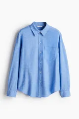Camisa celeste de tejido liviano en mezcla de lino y viscosa, con cuello clásico, botones al frente, canesú de doble capa con pliegue de caja en la espalda, mangas largas con puños abotonados, bolsillo superior abierto y bajo redondeado.