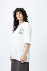 Remera color beige de corte amplio con mangas cortas y estampado de una postal con una palmera en el pecho.
