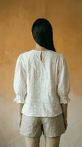 Blusa de color natural confeccionada en tejido 100% algodón. Presenta un cuerpo delantero fruncido tipo smock, vuelo peplum en la cintura y mangas globo 3/4 con volados en los puños.