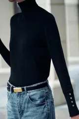 Jersey negro de punto liso con cuello alto, manga larga y detalle de botones dorados en los puños.