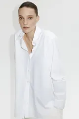 Camisa blanca de algodón con cuello clásico, abotonada adelante y corte holgado. Tiene mangas largas con puños sencillos y bajo redondeado con espalda algo más larga.