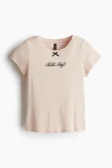 Remera rosa de algodón con cuello redondo y mangas cortas. Presenta un pequeño moño negro y el logo de Hello Kitty bordado en el pecho.