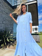 Vestido camisero largo celeste con rayas verticales blancas, cuello camisero, manga larga con puño y abotonadura frontal. Diseño escalonado con volantes en cada corte.