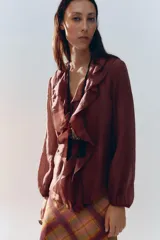 Camisa color crudo confeccionada en tejido con lyocell. Presenta escote en pico, mangas largas abullonadas con elástico en los puños, volantes frontales y cierre frontal con botones.