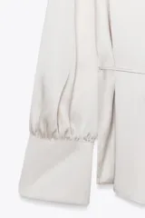 Camisa color blanco hueso de satén, con cuello solapa, escote en pico y mangas largas con puños abotonados.