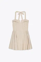 Vestido corto a cuadros beige y blanco, con cuello halter que se ata en la nuca, escote recto con abertura y falda acampanada. Tiene bolsillos laterales ocultos, espalda elastizada y botones decorativos en el frente.