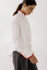 Camisa blanca de poplin con cuello mao, manga larga y cierre frontal con botones. Presenta detalles de crochet rojo en cuello, puños y bolsillo.