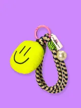 Llavero con aro rosado, dije de peluche amarillo con cara sonriente, cordón trenzado con estampado de pata de gallo, cordón trenzado verde y dije con forma de etiqueta con la marca She Mona y una perla sintética.