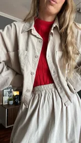 Camisa corta color beige de pana con cuello camisero, cierre frontal con botones, manga larga con puños elásticos, bolsillo de parche en el pecho y corte a la cintura con elástico.