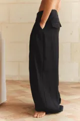 Pantalón negro de tiro alto con trabillas, doble pliegue, bolsillos laterales y tapetas en la espalda. Tiene pernera ancha y cierre frontal con cremallera y gancho metálico.