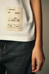 Camiseta blanca de algodón, corte recto, cuello redondo y manga corta. Tiene una etiqueta estampada en el frente.
