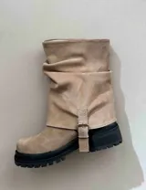 Bota de caña media color beige, con puntera cuadrada y suela track negra. Presenta una correa con hebilla metálica en el lateral.
