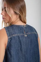 Blusa sin mangas de denim oscuro, con cuello redondo y costuras contrastantes en color marrón claro. Presenta un pequeño parche rectangular multicolor en la parte inferior frontal. El ruedo es deshilachado.