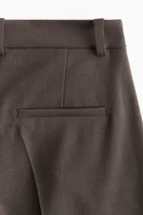 Pantalón sastrero marrón de corte holgado con tiro alto y cierre con corchete y botón ocultos. Piernas amplias con pinzas, pliegues adelante y atrás, bolsillos al bies y bolsillos decorativos atrás.