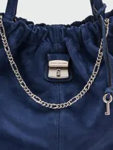 Bolso tipo hobo o bucket de gamuza marrón, con frunce en la parte superior y detalles de costura en contraste. Cuenta con una cadena dorada decorativa y un dije metálico con logo.