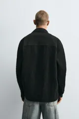 Camisa negra de denim de algodón con textura irregular, corte relaxed fit, cuello solapa, manga larga con puños abotonados y cierre frontal con botones.