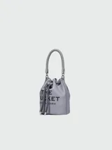 Bolso tipo bucket de cuero color gris con logo "The Bucket Marc Jacobs" estampado en negro. Tiene asa corta de cuerda trenzada y correa larga de cuero ajustable y removible.