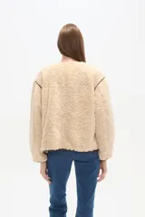 Campera de poliéster con textura de corderito en color beige, con cuello a la base y cierre frontal de botones. Presenta ribetes en contraste de color marrón oscuro en el cuello, la cartera, los hombros y en sus dos bolsillos frontales tipo parche.