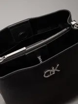 Cartera tipo saco Calvin Klein color negro, con correa ajustable para el hombro, compartimento central con cremallera, compartimento con cierre giratorio en la parte delantera, compartimento magnético en la parte trasera y herrajes plateados con el monograma de la marca.
