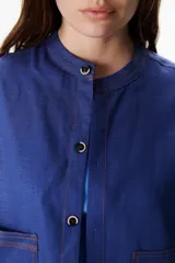 Chaqueta color crudo de lino con cuello redondo, manga tres cuartos, cierre frontal con botones y bolsillos de parche. Detalle de costuras en contraste color marrón.