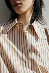 Camisa oversized de sarga color beige con rayas verticales marrones, cuello clásico, cierre frontal con botones ocultos, mangas largas con puños abotonados y bajo recto.