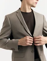 Traje de vestir color marrón topo, compuesto por saco de dos botones con solapa de muesca, dos bolsillos delanteros con solapa y un bolsillo superior. Pantalón de vestir con corte recto y bolsillos laterales. Incluye camisa blanca y corbata azul con lunares blancos.