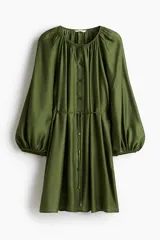 Vestido corto color verde oliva, de corte holgado, con cuello redondo fruncido, mangas globo largas con puños elásticos y botonadura frontal. Incluye cinturón fino para atar.
