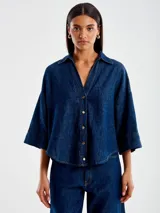 Camisa de denim azul con corte holgado, escote en V, cuello polo, mangas kimono y botones dorados.