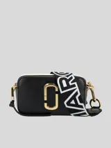 Bolso bandolera tipo cámara (camera bag) Marc Jacobs The Snapshot, color negro con detalles en blanco y herrajes dorados. Presenta el logo 'J' en metal dorado en el frente y una correa ancha de tela con el logo de la marca en blanco y negro.