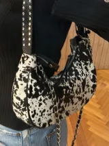 Cartera tipo shoulder bag con estampado animal print de vaca en tonos marrón y blanco, con correa de cuero y cadena metálica.
