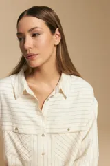 Camisa de manga larga color blanco hueso con finas rayas verticales en color oscuro. Presenta cuello camisero, botones frontales y dos bolsillos superpuestos en el pecho.
