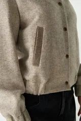 Campera estilo bomber de cuello redondo, color beige jaspeado, con cierre frontal de broches y bolsillos delanteros con ribete marrón.