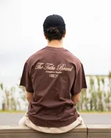 Remera marrón de corte oversize confeccionada en jersey de algodón, con estampa en la espalda con el texto "The Tribu Brand Creative Studio" en color crema y un diseño de tres líneas horizontales debajo.