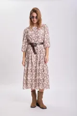 Vestido largo con estampado floral en tonos mostaza, marrón y blanco, con mangas largas abullonadas y falda con paneles. Cinturón marrón con tachas.
