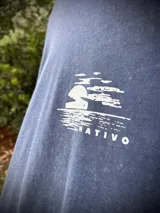 Remera oversize de algodón con efecto lavado (acid wash) en tono azul grisáceo. Presenta un estampado circular en la espalda con una palmera y texto que dice "SUNSET", "TROPICAL" y "PALM BEACH".