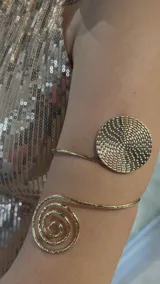Brazalete dorado para brazo, con diseño de espiral en un extremo y círculo con textura en el otro.