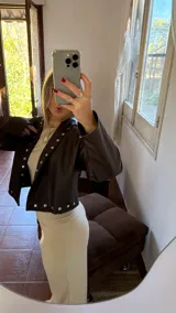 Campera corta de cuero sintético color marrón oscuro, con cuello solapa y detalles de tachas plateadas en el frente, hombros y puños. Presenta mangas amplias.