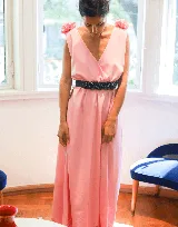 Vestido largo color rosa pálido, con escote en V cruzado, sin mangas y cinturón negro con apliques de flores. Presenta detalle de volados en los hombros y corte evasé.