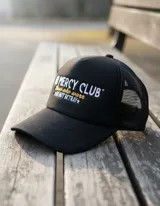 Gorra trucker negra con frente rígido y rejilla trasera. Bordado frontal con la frase "No Mercy Club" en blanco y amarillo.
