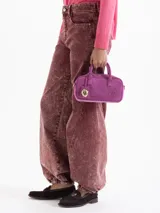 Cartera tipo baulito de cuero descarne vacuno en color fucsia, con diseño compacto y estructura rígida. Presenta doble asa de mano, logo de la marca grabado en relieve en el frente y un charm metálico en forma de vaquita de San Antonio colgando de una de las asas.