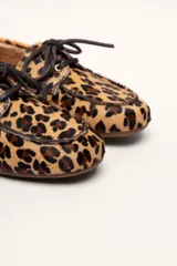 Mocasín tipo boat shoe con estampado de vaca en tonos marrón y blanco, confeccionado en cuero. Cuenta con cordones de cuero marrón oscuro y suela de goma.