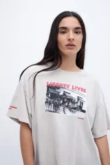 Remera de algodón color beige claro con efecto lavado, corte boxy y manga corta. Estampado frontal con una fotografía en blanco y negro de un local con el texto "LIBERTY LIVES" en rojo. Pequeño texto "CHARM" en la manga izquierda.